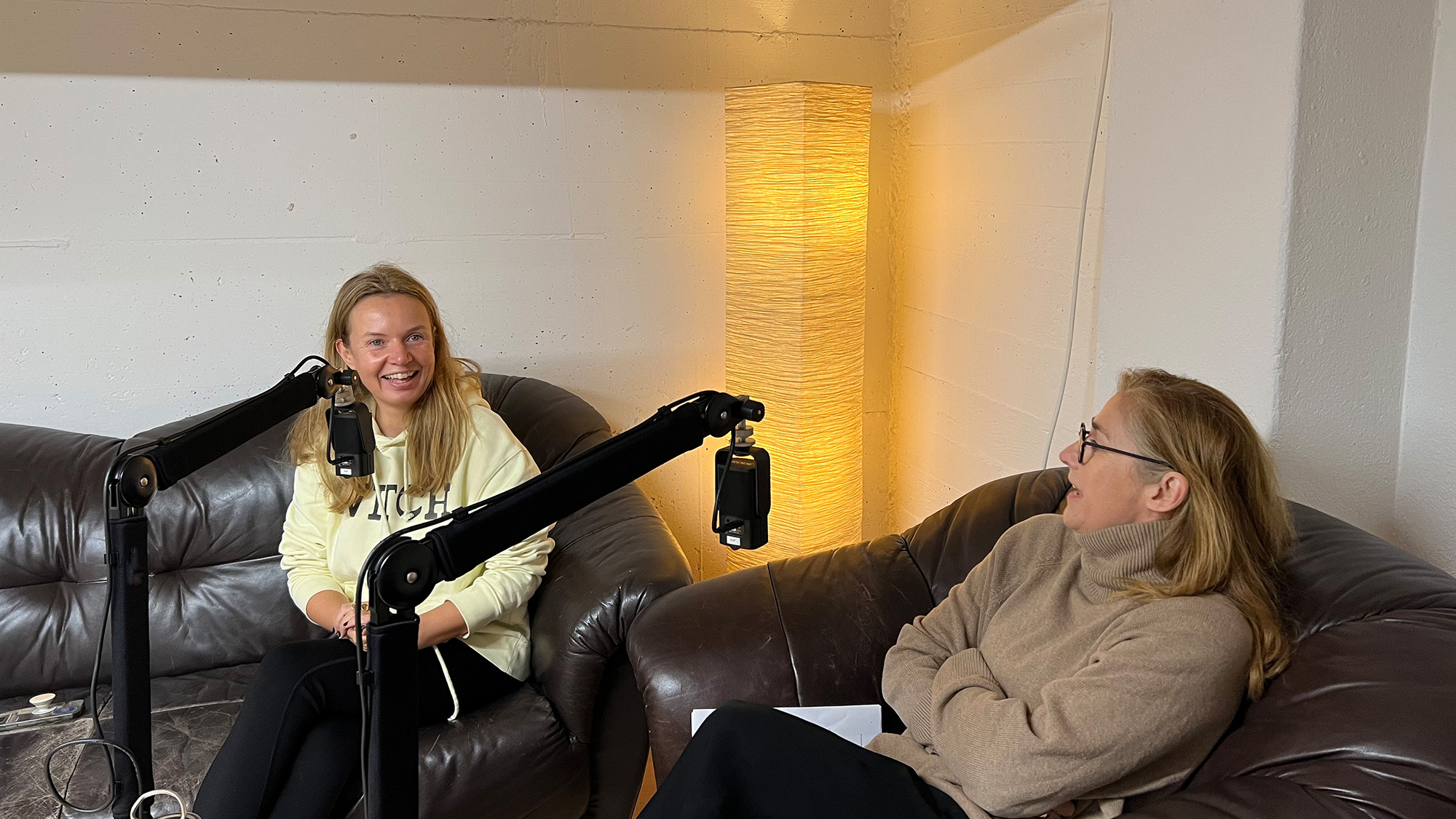 Bildbeschreibung: Foto von der Podcast-Aufzeichnung. Eine Person mit blonden langen Haaren und pastellgelbem Pullover sitzt aufrecht auf einem dunklen Ledersofa und blickt lächelnd in die Kamera, eine zweite Person mit braunem Pullover, Brille und halblangen hellbraunen Haaren sitzt rechts davon zurückgelehnt und mit verschränkten Armen in einem Ledersessel, sie blickt ins Leere und spricht halb der ersten Person zugewandt. Im Vordergrund zwei Mikrofonarme, im Hintergrund sieht man die Ecke eines Raumes mit tiefer Decke, darin steht ein Stehlampe aus Stoff und verströmt warmes Licht.