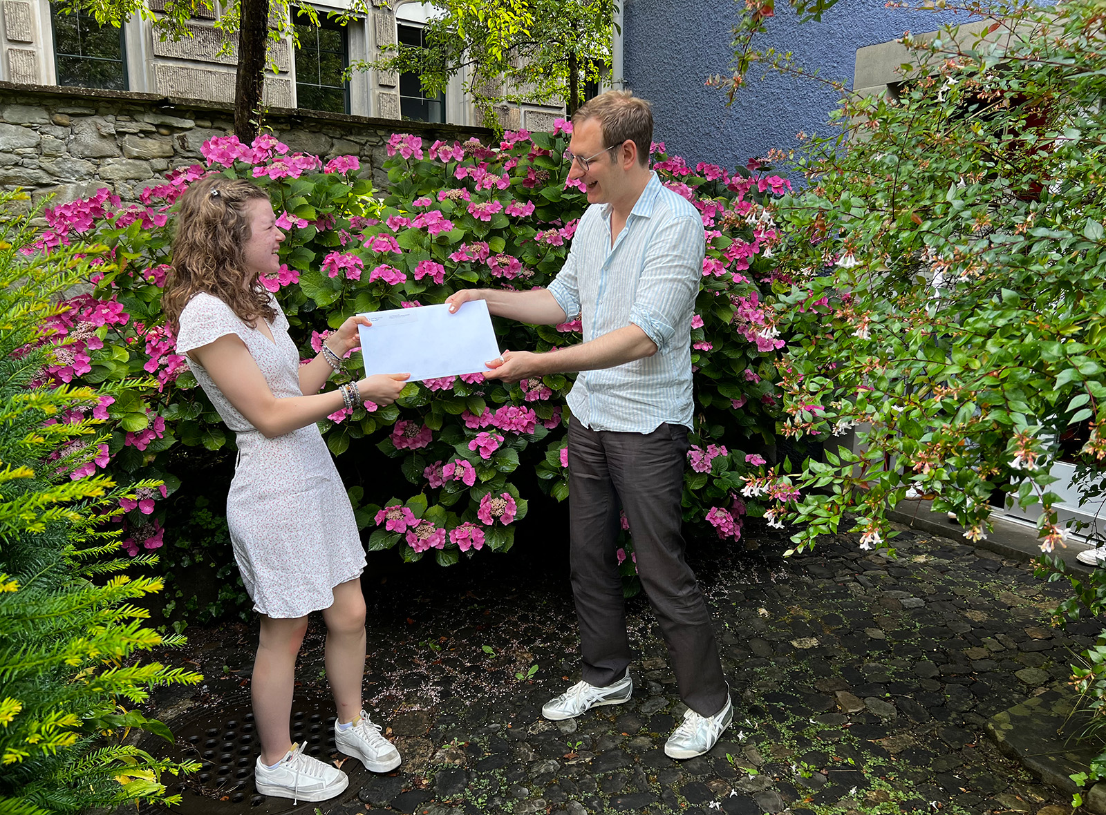 Eine junge Frau im hellen Sommerkleid steht in einem Garten vor einem Busch voller Blumen und nimmt von einem Mann in hellem Sommerhemd und dunklen Anzughosen ein Couvert entgegen.