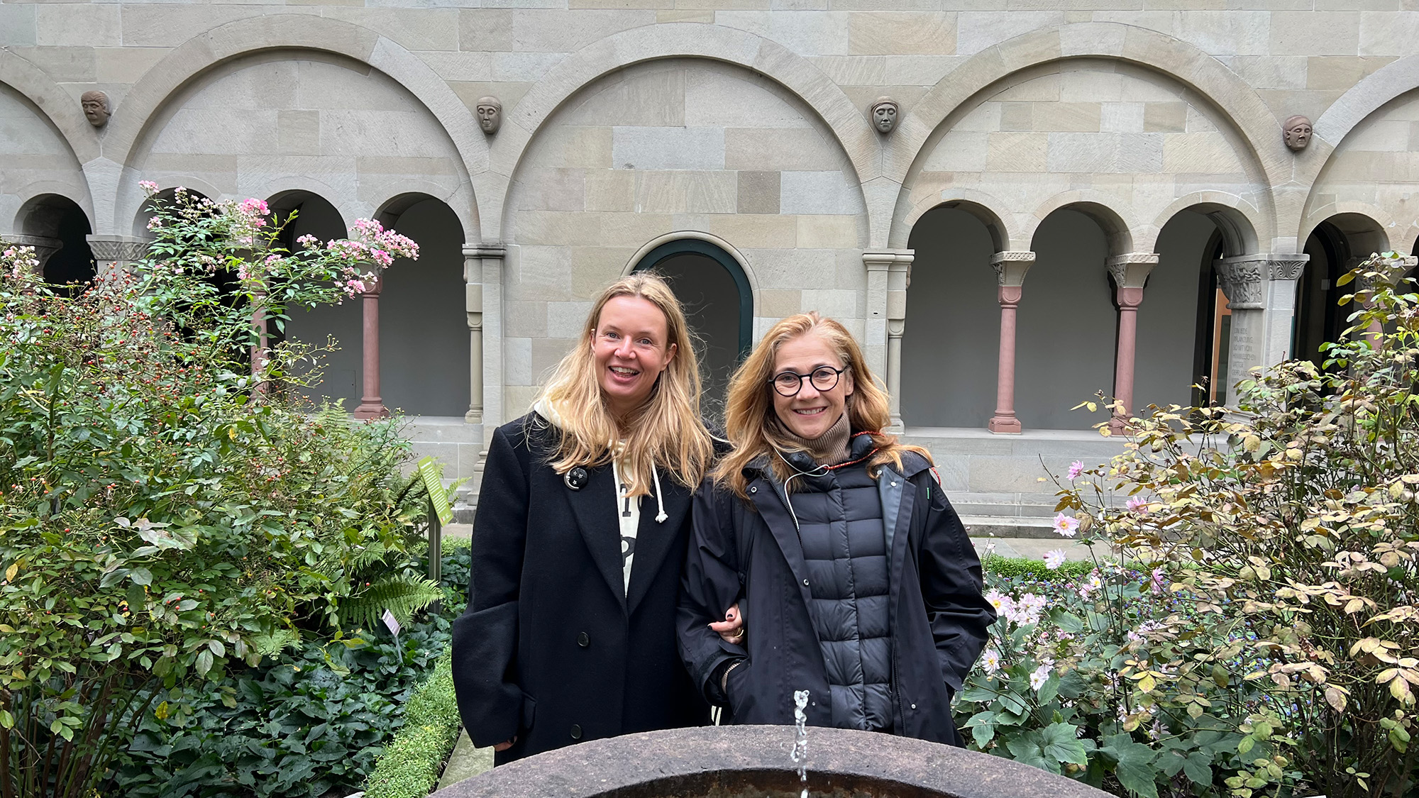 Bildbeschreibung: Foto von dem Treffen im Vorfeld der Podcast-Aufzeichnung. Zwei Personen mit langen, offenen Haaren stehen eingehakt mit Mänteln hinter einem kleinen Springbrunnen und lächeln in die Kamera. Neben Ihnen sind lockere, hohe Büsche zu sehen. Im Hintergrund die Säulen und Bögen des Kreuzgangs mit grauen Steinen und dunkelroten Säulen.